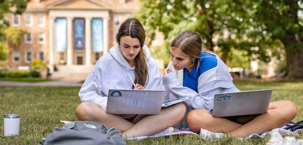 Students on their laptops sitting on the grass of Polk Place. South Building is visible behind them in the background.