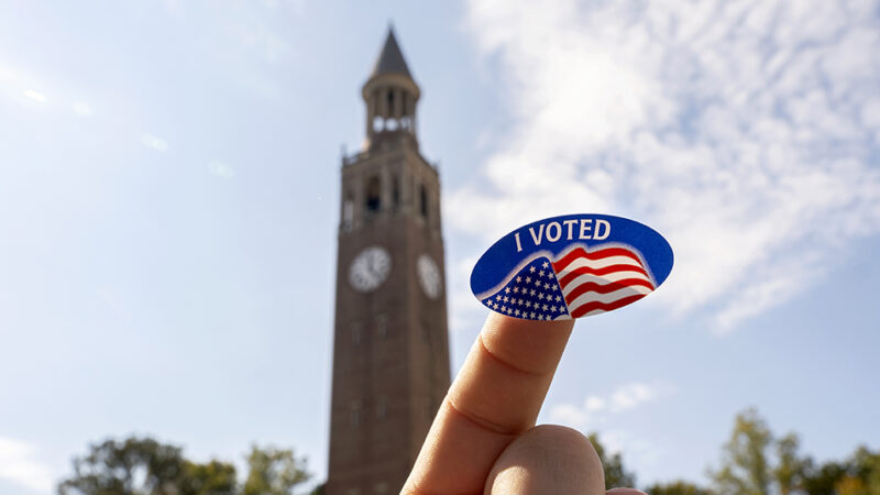 Photo showing voter sticker with the UNC Bell Tower in the background. On a clear sunny day with blue clouds.